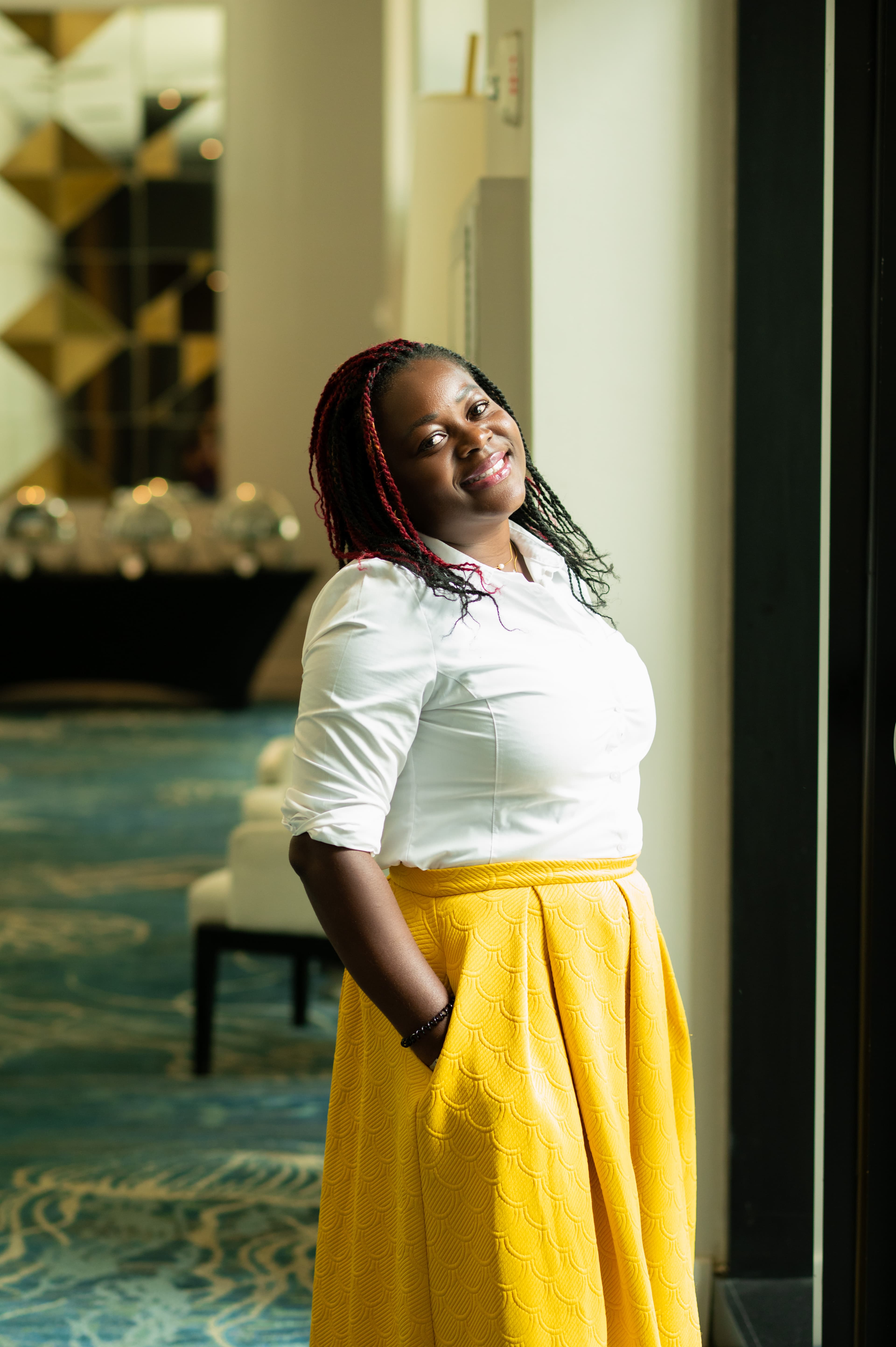 Anwin Mbah standing by a window in a white shirt and yellow skirt, smiling warmly